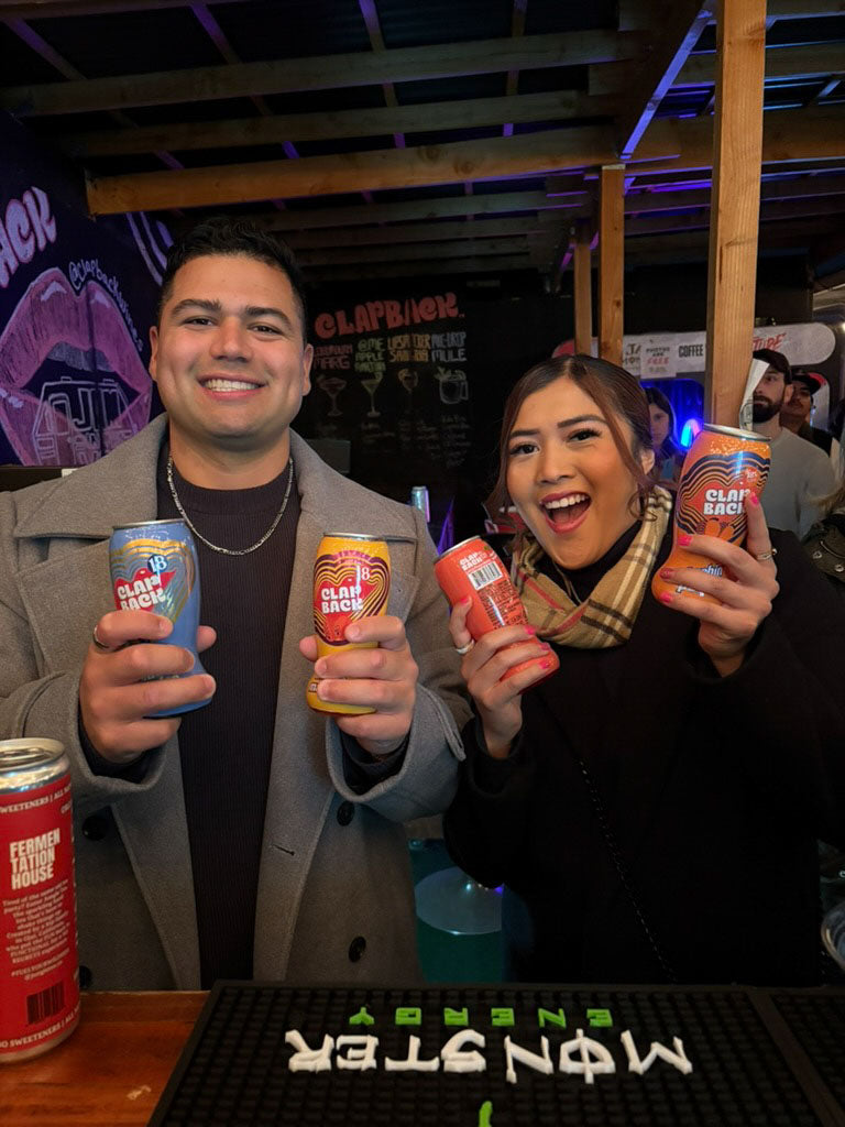 Two people holding cans of ClapBack grape wine in a bar setting