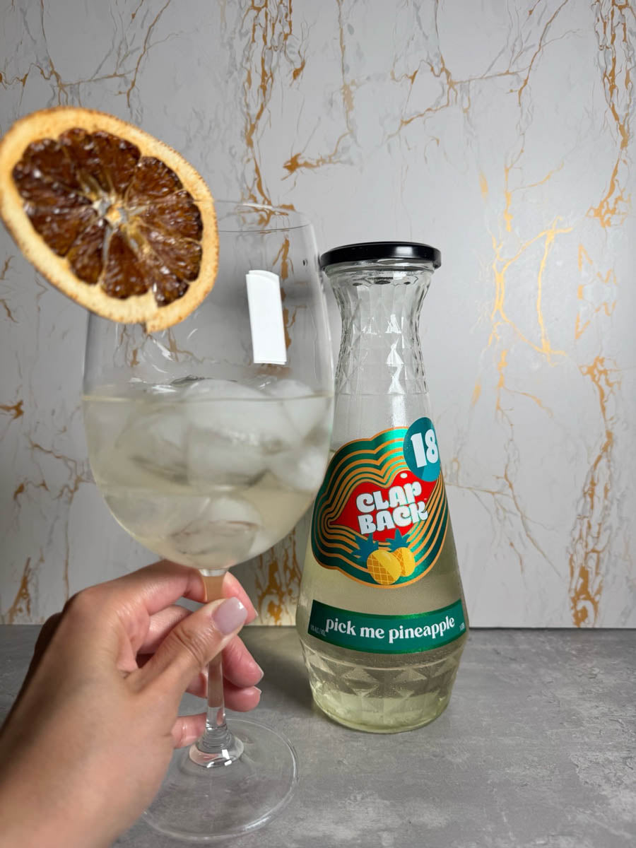 Glass of wine with a dried orange slice, Clap Back bottle, and hand holding the glass against a marble background.