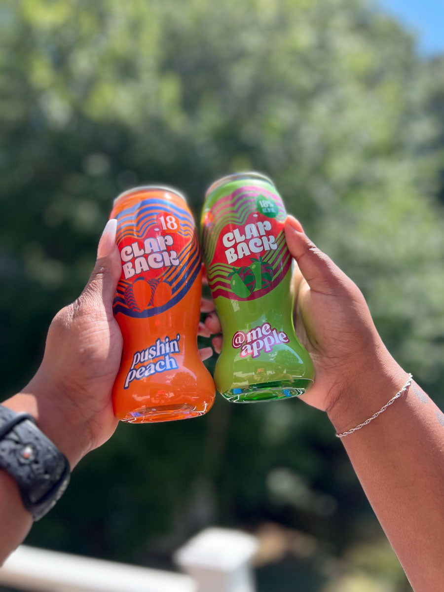 Two hands holding an orange and a green can of Clap Back soda against a blurred green outdoor background.